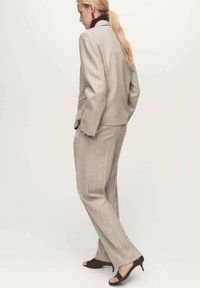Un costume gris clair à rayures pinstripe comprend un blazer ajusté et un pantalon droit, associé à des sandales à talons ouverts de couleur marron foncé.