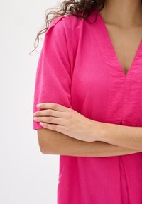 Fukcija ružičasti top s kratkim rukavima, dizajn s V izrezom i teksturiranom tkaninom. Značajni detalji uključuju šavove duž prednje strane i obrnute manžete.