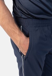Marineblaue Sporthosen mit elastischem Bund, seitlichen Reißverschlusstaschen und weißen Paspelierungen an den Kanten, gefertigt aus einem glatten, leichten Stoff.