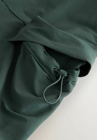 Material verde închis cu o textură netedă, având o manșetă strânsă și detalii cu șnur elastic pentru ajustare.