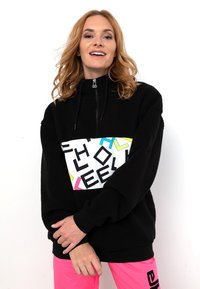 Schwarzer Fleece-Pullover mit einem Halb-Reißverschluss, der ein buntes Grafikpaneel mit Buchstaben in Rosa, Blau und Gelbakzenten zeigt.