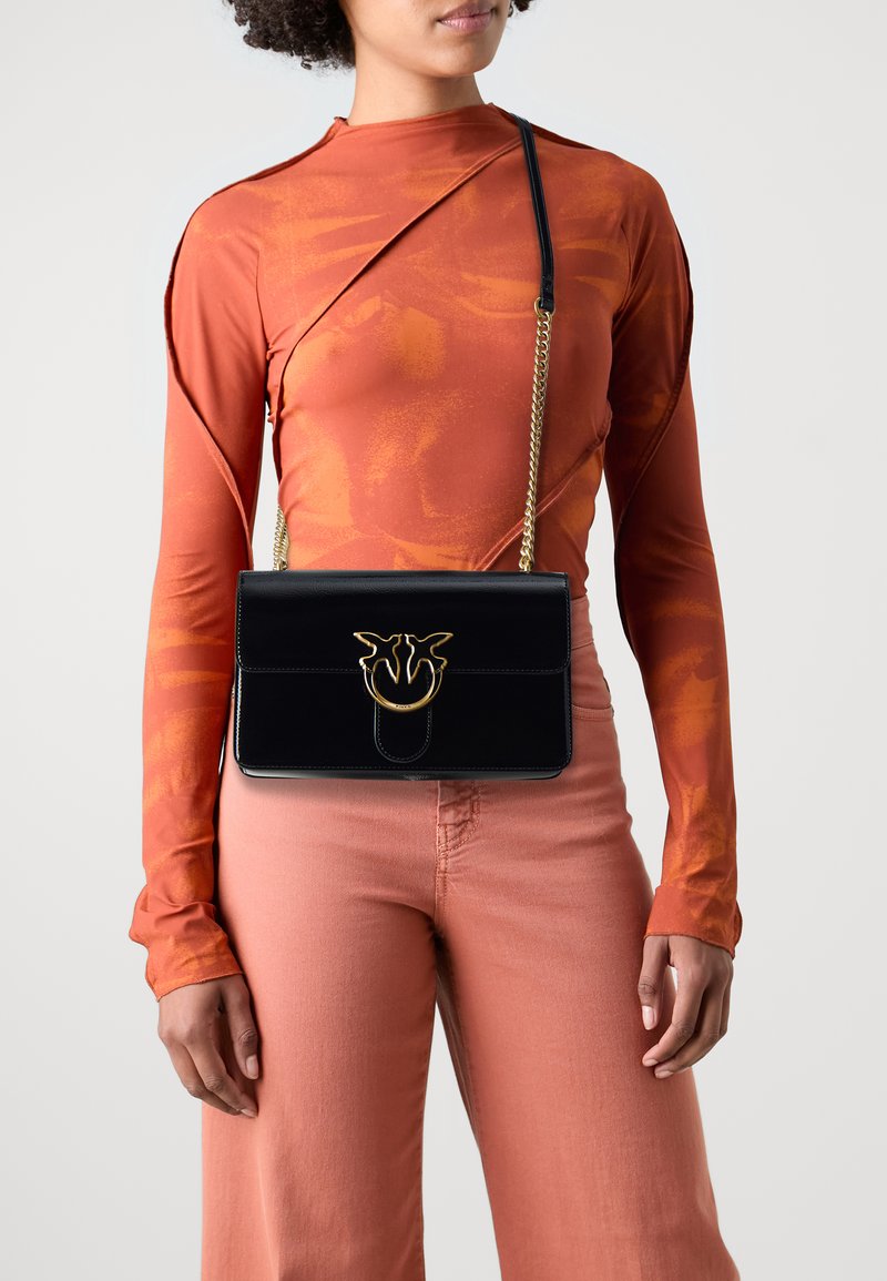 Černá kožená crossbody taška s módním zlatým kováním, kompaktně obdélníkového tvaru, s řetízkem na nošení. Nositelná s oranžovým topem a růžovými kalhotami.