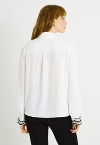 Blusa blanca de manga larga con textura arrugada, con detalles de encaje negro en los puños y un bajo recto. Diseño de cuello clásico.