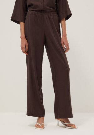 Female model wearing dark brown wide-leg pants with elastic waist paired with white heeled sandals against a plain light background.