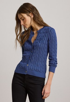 Frau, die eine blaue Strickjacke mit Zopfmuster und goldenen Knöpfen sowie schwarze Hosen trägt, posiert vor einem einfarbigen hellgrauen Hintergrund.