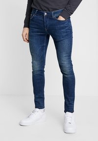 Slim-fit, tmavě modré denimové džíny s vybledlým designem, se zipem a knoflíkovým zapínáním, pěti kapsami, nošené s bílými teniskami.