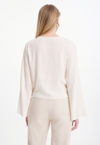 Top de un beige claro, holgado, con mangas acampanadas largas y un escote amplio, fabricado con una tela suave y texturizada; el dobladillo se sitúa a la altura de la cintura.