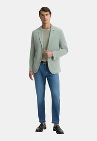 Blazer verde chiaro, camicia beige, jeans blu e scarpe in pelle nere. Il blazer presenta due tasche frontali e una chiusura con un bottone.