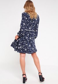 Dunkelblaues Kleid mit langen Ärmeln, das ein himmlisches Muster aus Sternen und planetarischen Symbolen zeigt, sowie eine taillierte Silhouette mit einem weißen Gürtel.