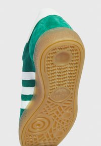 Grön mockaskärpa med vita ränder och en gummigummi-sula. Har en rundad tå och ett strukturerat mönster på undersidan.