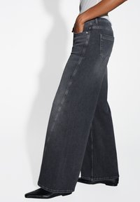 Wijdvallende spijkerbroek in donkergrijs denim. Voorzien van voor- en achterzakken, subtiele stiksels en een rechte, ontspannen pasvorm. Schoeisel is zwart met een spitse neus.