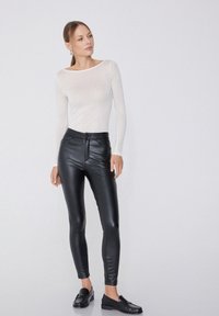 Schwarze, fauxleder Leggings mit hohem Bund, ausgestattet mit einem Reißverschluss und Knopfverschluss vorne. Kombiniert mit einem leichten, langärmeligen Oberteil.