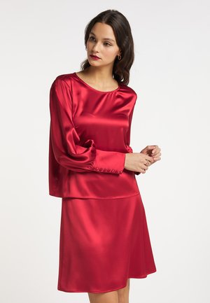 Frau trägt eine glänzende rote Satinhbluse mit langen Ärmeln und passenden roten Satinhrock, mit losem dunklem Haar und dezentem Make-up.