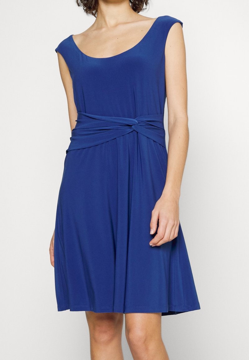 Kvinna klädd i en ärmlös blå klänning som når knäna, med rundad halsringning och knytning i midjan.