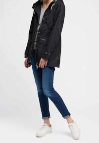 Parka negra con capucha y cordones, dos bolsillos frontales y dobladillo curvado, combinada con vaqueros azules con dobladillo enrollado y zapatillas blancas.