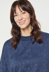 Tengerészkék pulóver, puha, texturált felülettel, széles kerek nyakkal és laza fazonnal. Nincsenek látható minták vagy fém részletek.