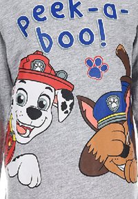 Grijze T-shirt met cartoonhonden, één in een rode brandweerpet en één in een blauwe politiepet, met de tekst "Boekeltje!" in blauw en rood.