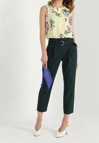 Blusa senza maniche gialla a fiori abbinata a pantaloni verde scuro e tacchi bianchi. Tiene in mano una pochette blu. L'outfit presenta una vita cintrata.