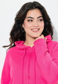 Sweat à capuche zippé fuchsia avec une texture douce, cordon de serrage et fermeture éclair complète. Le modèle tient le tissu au niveau du cou, mettant en avant son ajustement.