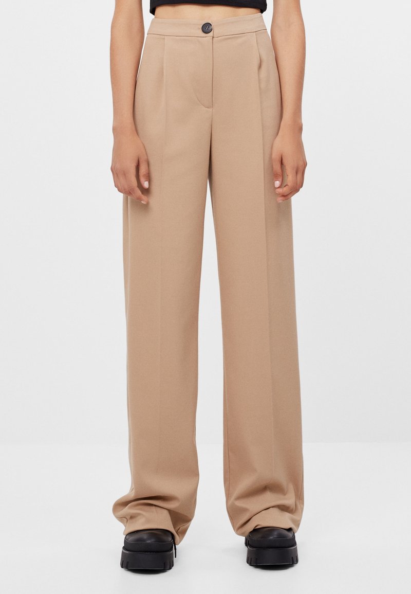 Pantalon beige à jambes larges, taille haute, fermeture par un seul bouton, plis sur le devant, porté avec des chaussures noires épaisses sur un fond blanc uni.