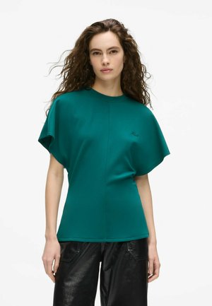 Femme aux cheveux bouclés portant un haut à manches courtes couleur sarcelle et un pantalon noir, debout devant un fond blanc.