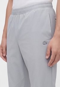 Pantalon gris clair à taille élastique avec poches latérales et petit logo crocodile sur la cuisse droite, porté par une personne en haut blanc.