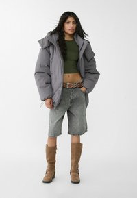 Graue Pufferjacke, grünes Cropped-Top, graue Jeansshorts, braune kniehohe Stiefel. Auffällige Details: Übergroße Passform, Gürtel mit metallischen Akzenten.