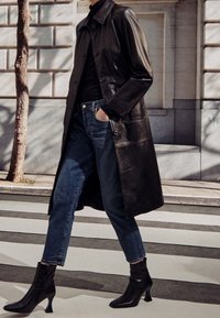 Lång svart läderrock med knäppningar, bärs över en svart topp och blå jeans. Avslutas med svarta ankelstövlar och en slank silhuett.