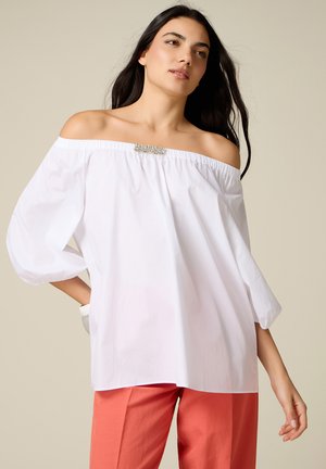 Frau mit langen dunklen Haaren, die eine weiße, schulterfreie Bluse mit Perlendetail und korallenrote, weite Hosen vor einem beigen Hintergrund trägt.