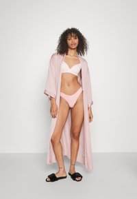Svijetloružičasti kimono ogrtač, lepršavog dizajna, otvorenog prednjeg dijela, nosi se preko odgovarajućeg ružičastog grudnjaka i bikinija. Crne plišane sandale na široku petu. Minimalistička pozadina.