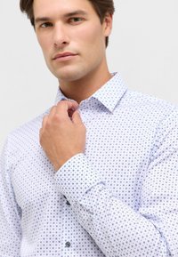 Camicia bianca con piccoli motivi circolari blu. Presenta un colletto a punta e maniche lunghe, mostrando una texture di cotone liscia.