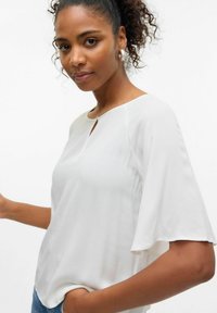 Kvinde med krøllet hår iført en hvid bluse med flagrende ærmer og lille nøglehulskrave, poserer med den ene hånd i jeanslommen.