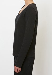 Person wearing a fitted black long-sleeve V-neck shirt and black pants, standing in profile against a plain white background.