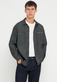 Camicia a righe grigia con bottoni, due tasche sul petto, logo ricamato in oro e una vestibilità comoda, indossata sopra una t-shirt bianca e jeans scuri.
