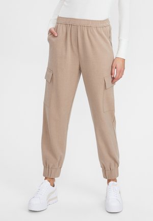 Beżowe spodnie typu jogger cargo z elastycznym pasem i mankietami, noszone z białymi trampkami i białą bluzką z długim rękawem.