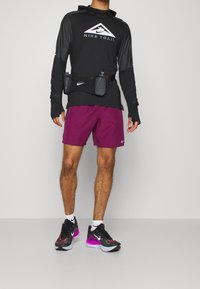 Haut à manches longues noir Nike Trail, shorts violets, et baskets noires et rouges avec une semelle grise. Comprend une ceinture d'hydratation noire avec deux bouteilles.