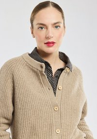 Beige gebreide cardigan met een ribbeltextuur, voorzien van een funnelsluiting, houten knopen en een gelaagd geruit shirt eronder.