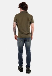 Polo de color verde oliva con mangas cortas, ajuste slim y detalles negros, combinado con jeans ajustados de color azul y zapatillas negras.