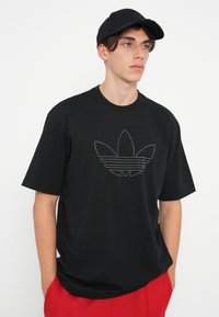 Melna īsām piedurknēm T-kreklu ar atstarojamā trīs lapu logotipa dizainu priekšpusē. Kombinēts ar sarkanām šortiem un melnu cepuri.