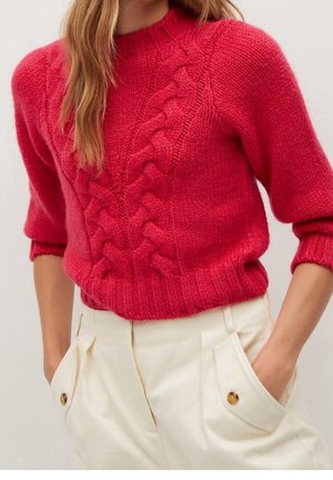 Femme portant un pull en maille torsadée rouge vif à manches bouffantes et un pantalon crème taille haute avec poches à boutons, les mains dans les poches.