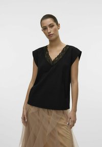 Top nero a V con pizzo, maniche corte e vestibilità rilassata, indossato con una gonna di tulle leggera color beige chiaro a strati e trasparente.
