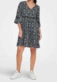 Žena v šatech po kolena s černobílým vzorem a rukávy po lokty, v bílých teniskách, stojící na pozadí bez vzoru.