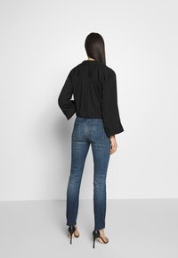 Černá blůza s širokými rukávy a záhybem na zádech, spárovaná se středně modrými džíny slim-fit. Silueta zvýrazněná vysokými podpatky.