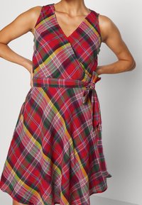 Červené a zelené kostkované bezramínkové zavinovací šaty s výstřihem do V a vázaným pasem. Vyrobené z lehké látky, splývavá sukně má volný střih.