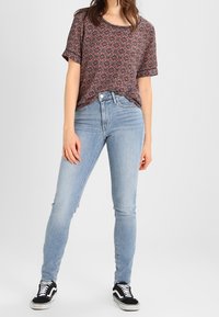 Camisa de manga corta con estampado en tonos oscuros, combinada con jeans ajustados de color azul claro. La lleva puesta con zapatillas negras y blancas. Look casual.