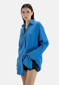 Camicia oversize in cotone blu con tessuto texturizzato e a coste, chiusura a bottoni e maniche lunghe. Abbinata a pantaloni shorts in denim neri.