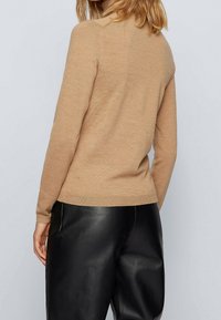 Suéter de punto beige con mangas largas y escote redondo. Presenta un dobladillo y puños acanalados, llevado sobre pantalones de cuero negros.