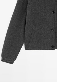 Gilet en tricot gris foncé avec une finition texturée, doté de boutons noirs et d'une poche latérale. Les poignets côtelés ajoutent une touche d'élégance.