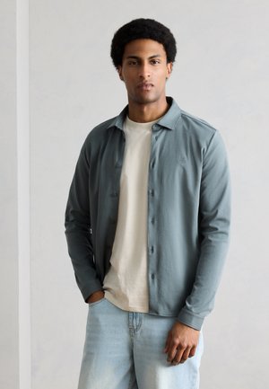 Νέος άνδρας με σγουρά μαλλιά, φορούσε μια τιρκουάζ πουκάμισο με κουμπιά πάνω από ένα μπεζ T-shirt και ανοιχτόχρωμα μπλε τζιν, στέκεται μπροστά σε ένα απλό ανοιχτό γκρι φόντο.