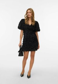 Melna ziedu kleita ar teksturētām rožu aplikācijām, īsām pūkainām piedurknēm un V-veida izgriezumu, kombinēta ar mazu melnu somiņu un smailiem augstpapēžu apaviem.